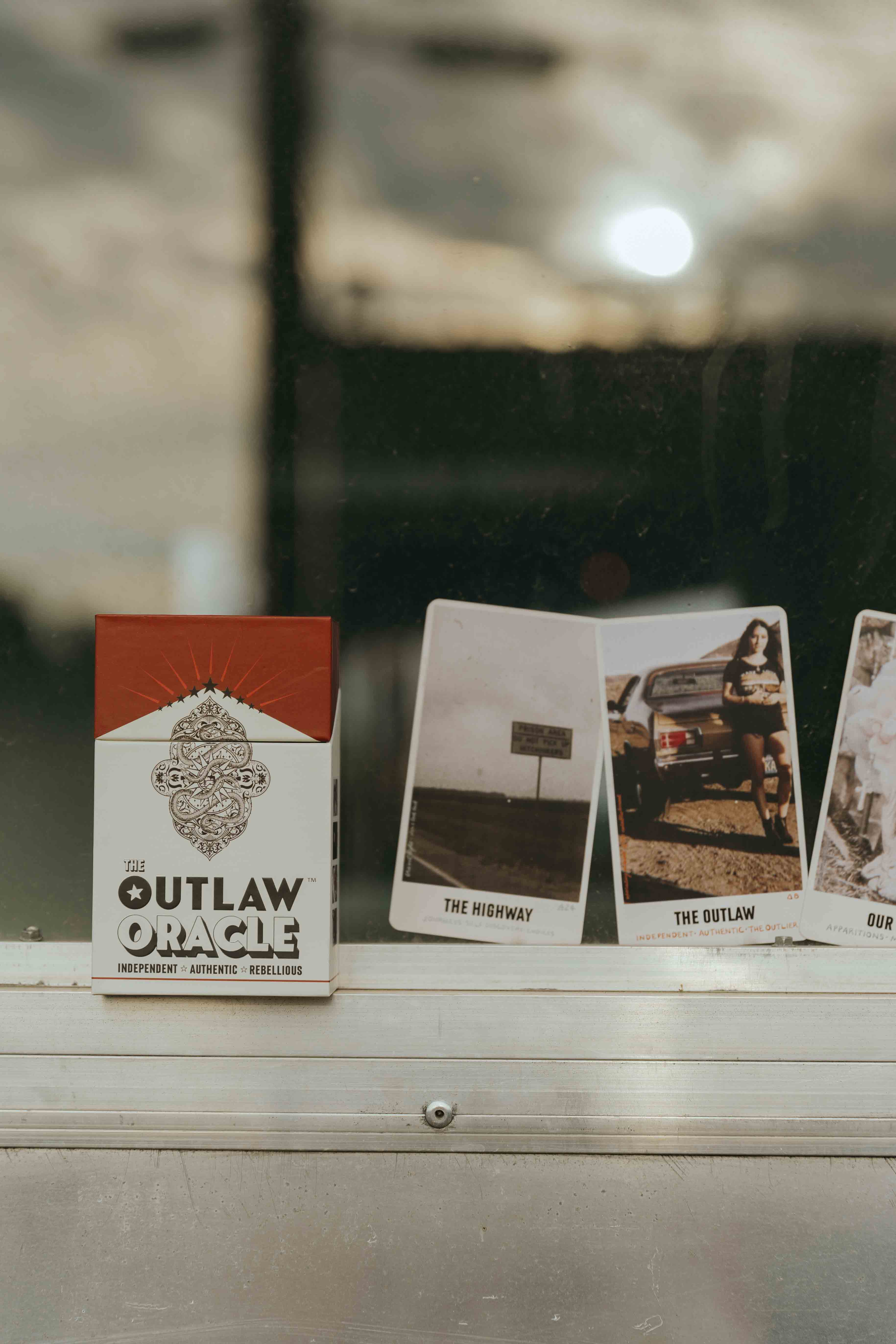 The Outlaw Oracle card deck box, with three of the cards featured within the deck - including The Highway card, The Outlaw Card and the Our Lady card.