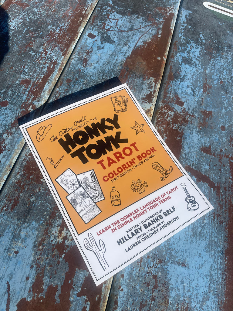 The Outlaw Oracle's Honky Tonk Tarot Coloring Book is laid across an old blue rusty picnic table.  It's bright yellow cover beckons you to look closer, seeing that this is a rare and unusual find.  A Honky Tonk take on the traditional tarot.  How novel.  